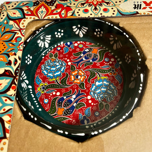 A hand-painted green ceramic ashtray with a vibrant red background and traditional floral motifs in blue, green, orange, and white, resting on a brown cardboard insert.