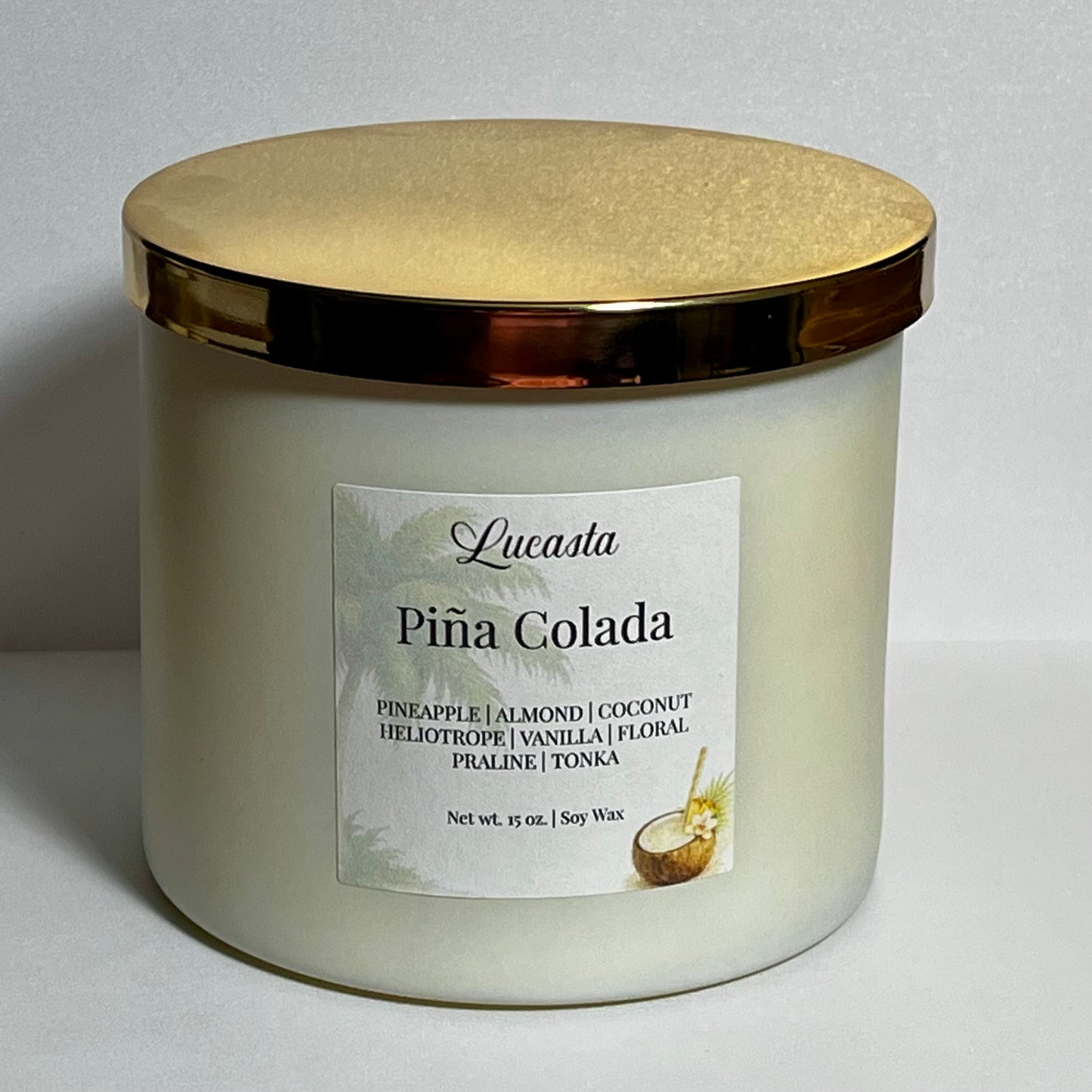 Close-up of Lucasta Piña Colada candle jar with gold lid and scent notes label reading pineapple, almond, coconut, heliotrope, vanilla, floral, praline, tonka