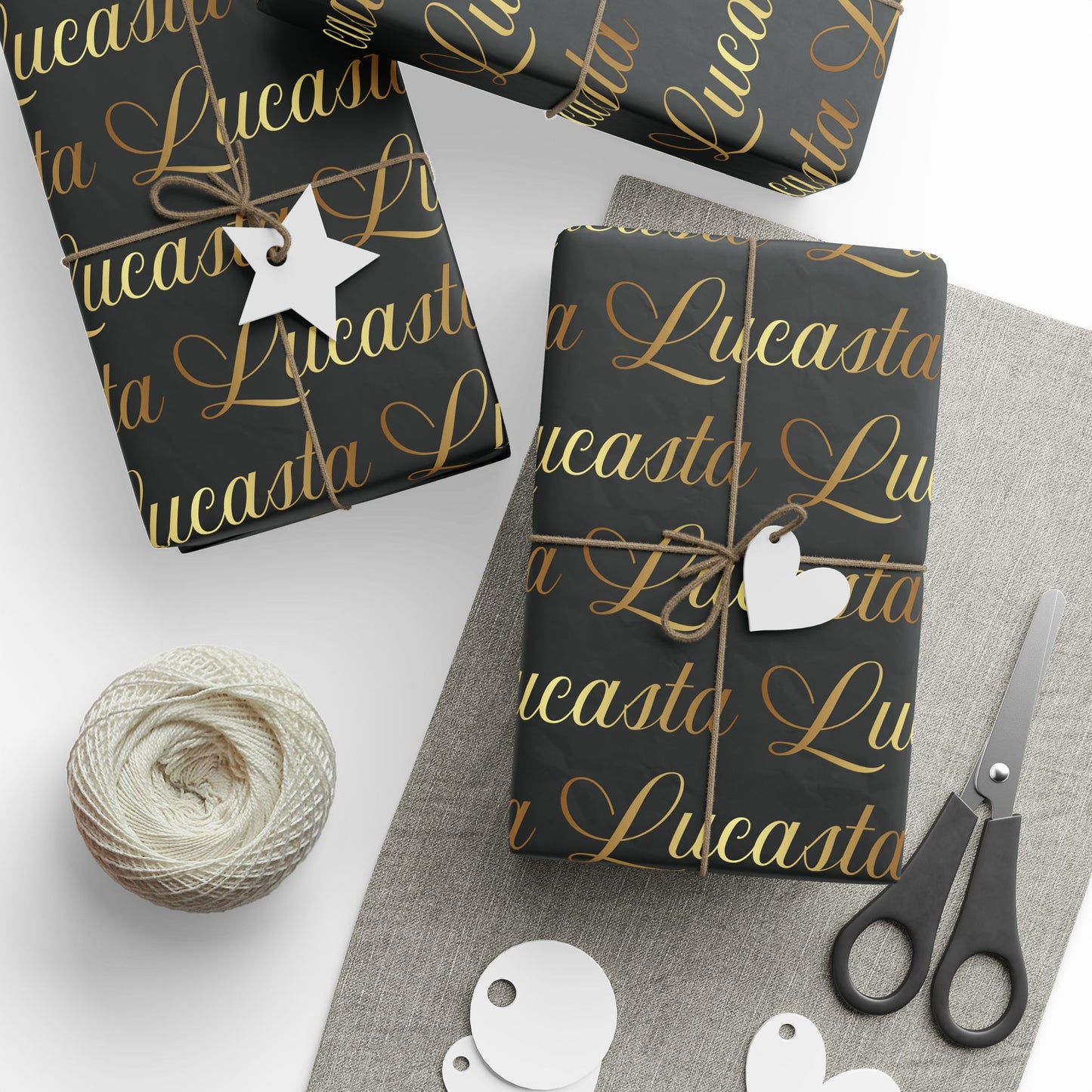 Lucasta black gift wrap with gold text, decorative tags, scissors, and yarn styled on a white surface