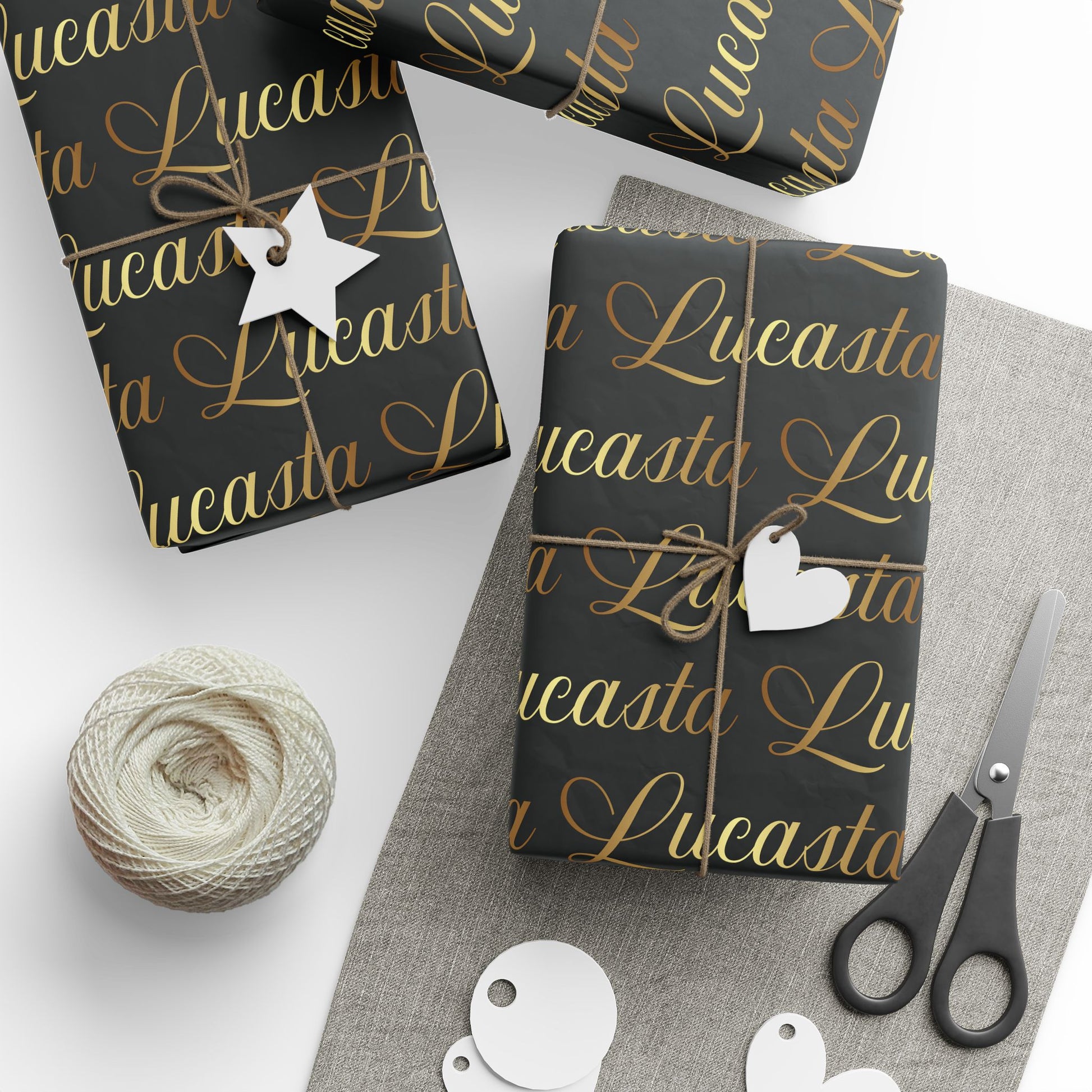 Lucasta black gift wrap with gold text, decorative tags, scissors, and yarn styled on a white surface