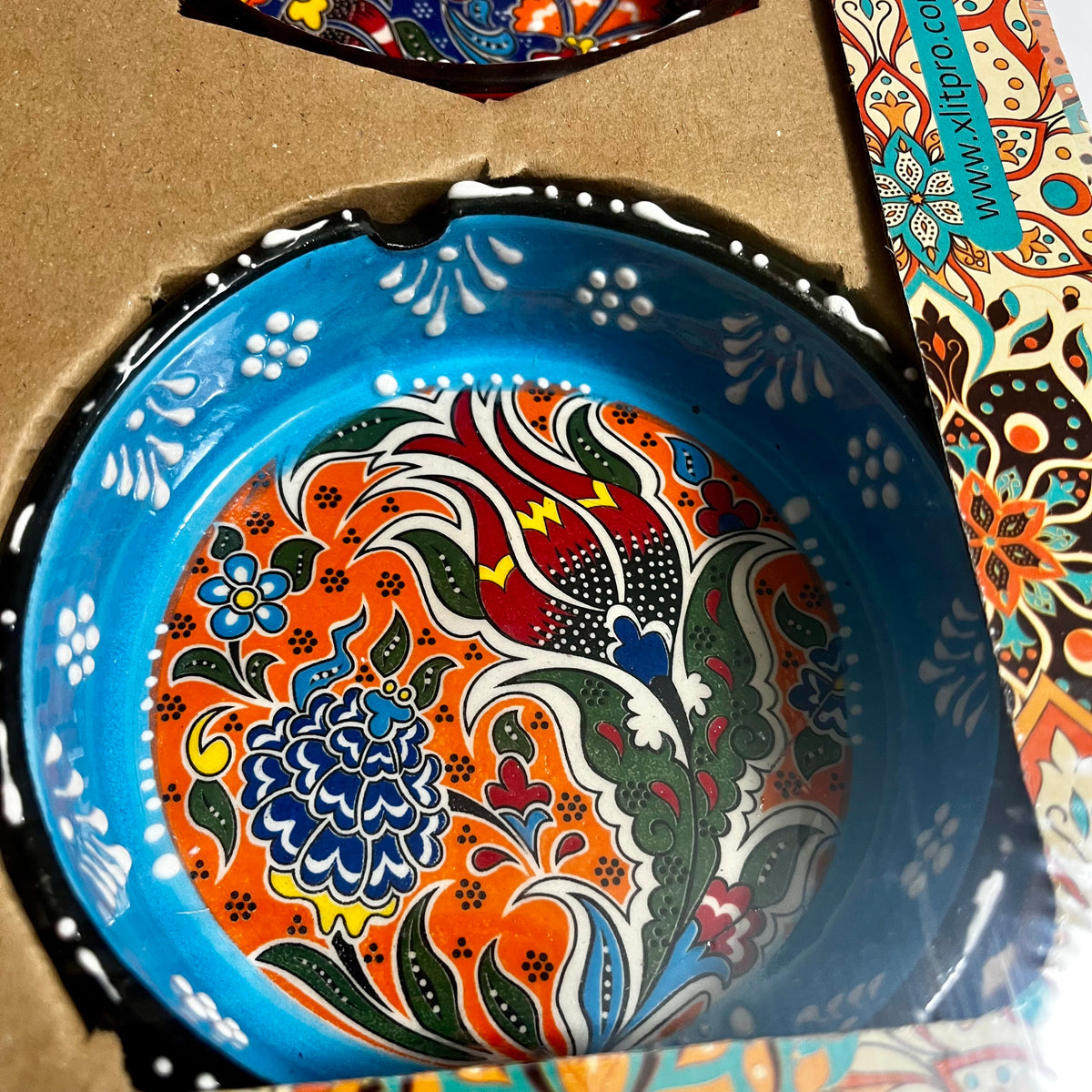 A close-up of a round ceramic ashtray with a vibrant hand-painted floral pattern, nestled inside its cardboard packaging. The plate features a bright blue rim and a multicolor interior with orange, red, green, blue, and white designs.