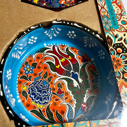 A close-up of a round ceramic ashtray with a vibrant hand-painted floral pattern, nestled inside its cardboard packaging. The plate features a bright blue rim and a multicolor interior with orange, red, green, blue, and white designs.