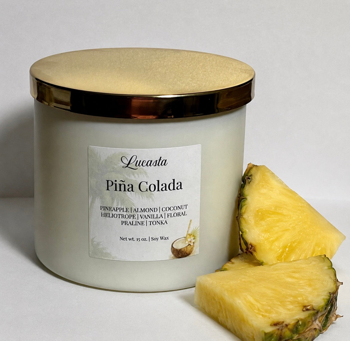 Lucasta Piña Colada soy candle in a white frosted jar with a gold lid, styled next to fresh pineapple slices on a white background