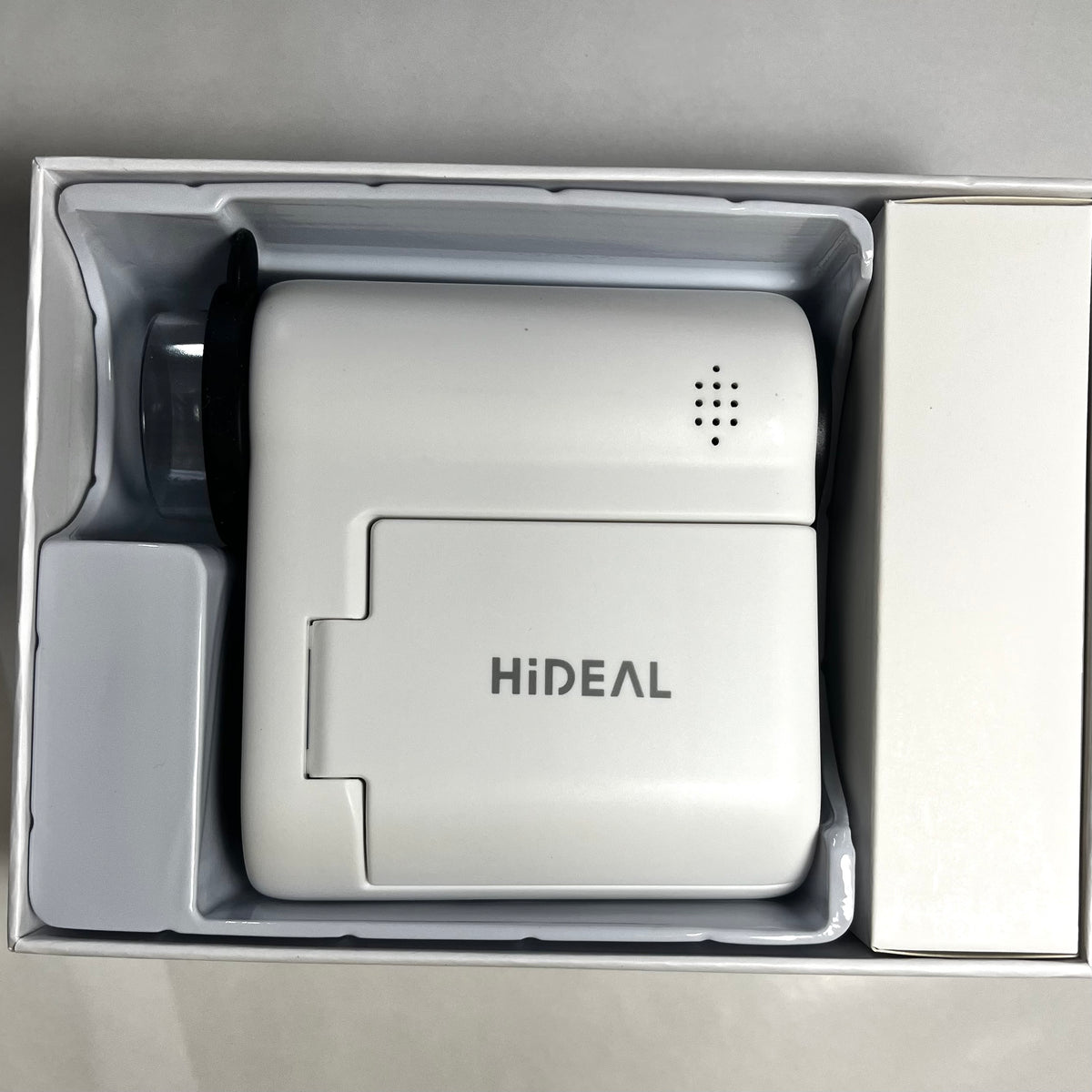 A white HiDEAL electronic microscope, shown inside its white retail packaging tray. The device features a hinged cover with the 'HiDEAL' brand name printed on it, a small speaker grille, and a black lens cover on the side.