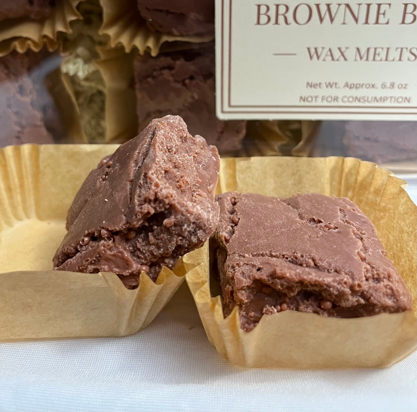 Two Lucasta Brownie Bites wax melts in kraft liners showing rich chocolate interior texture, with branded packaging in background