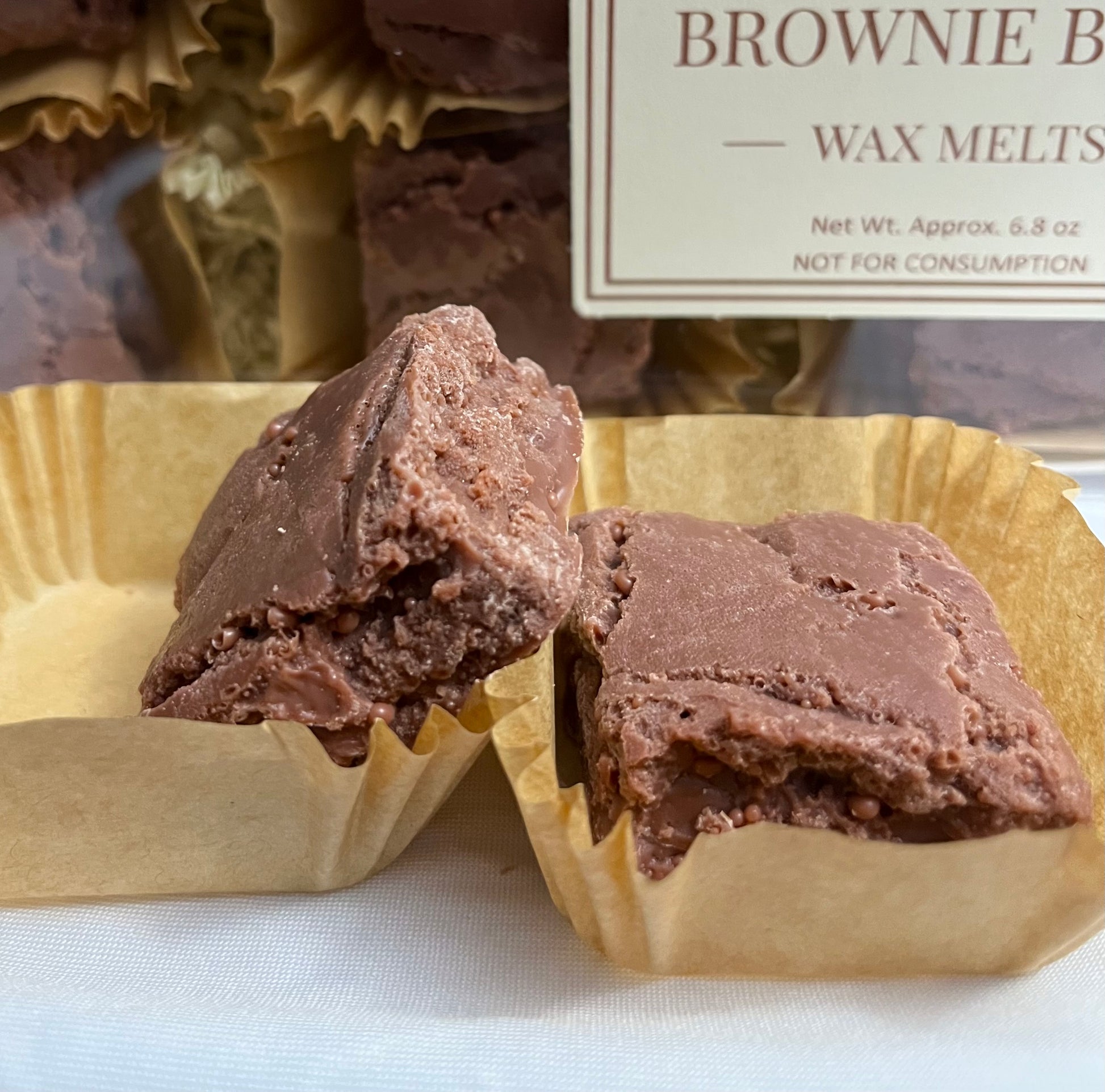 Two Lucasta Brownie Bites wax melts in kraft liners showing rich chocolate interior texture, with branded packaging in background