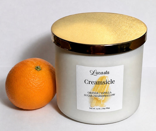Lucasta Creamsicle soy candle in a white frosted jar with a gold lid, styled next to a fresh orange on a white background