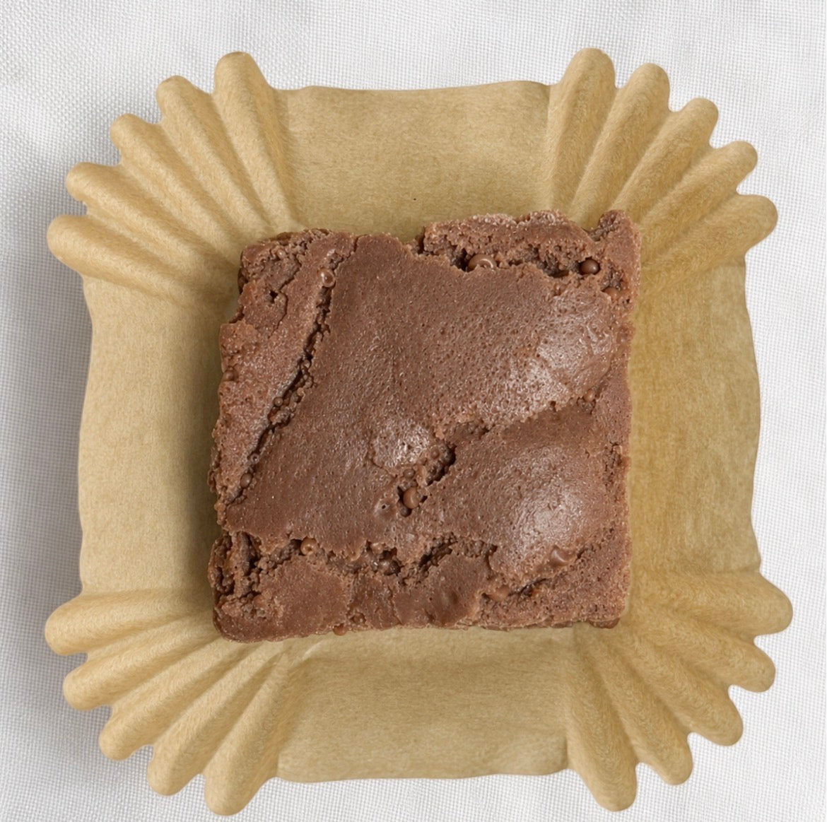 Close-up top view of a single Lucasta Brownie Bites wax melt in a kraft paper liner, showing realistic fudgy brownie texture