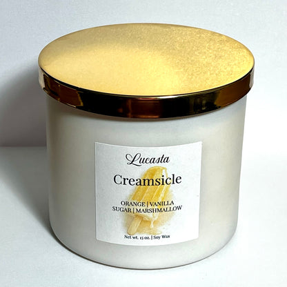 Close-up of Lucasta Creamsicle candle jar with gold lid and scent notes label reading orange, vanilla, sugar, marshmallow