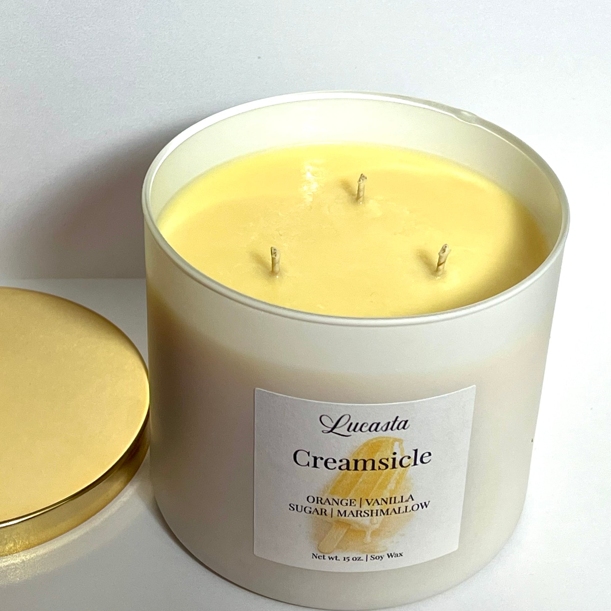 Lucasta Creamsicle triple-wick soy candle with gold lid resting beside it, showing the pale yellow wax and three wicks