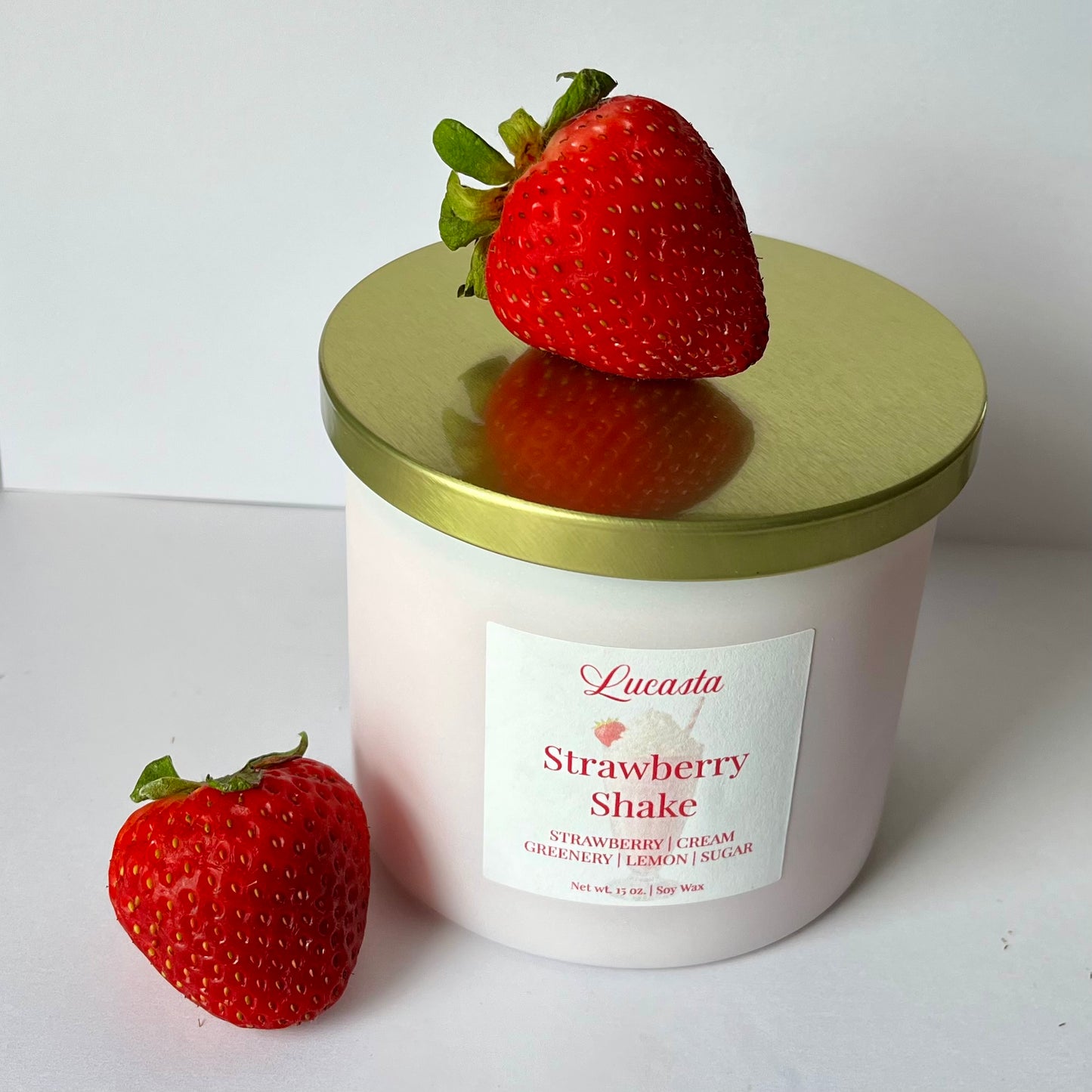 Lucasta Candles Strawberry Shake soy candle in a soft pink jar with a gold lid, styled with fresh strawberries on a white surface